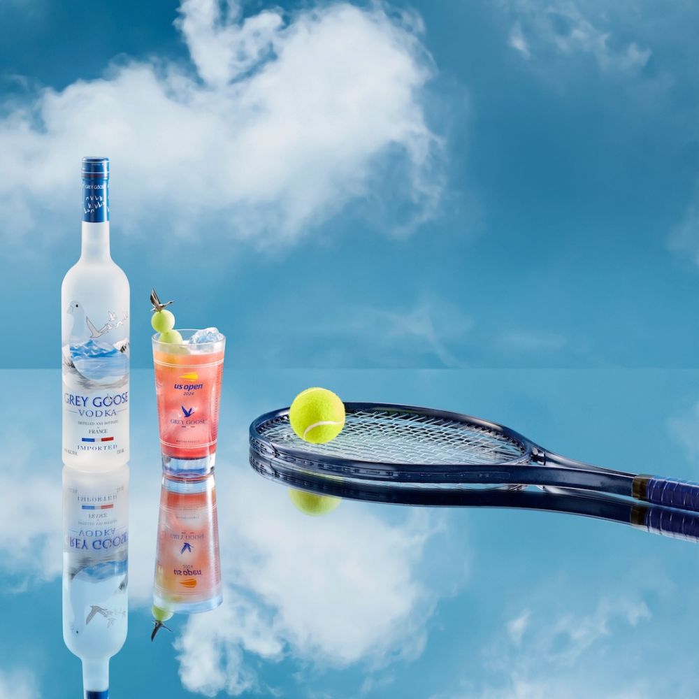 Grey Goose vodka bottle and US Open cocktail with tennis ball and racket against a cloudy blue sky background on a reflective surface.