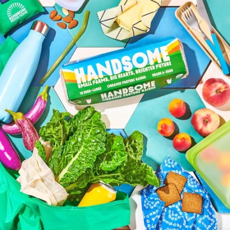 Overhead shot of organic produce, vegetables, fruit, and snacks on a colorful tile surface with green shopping bag and reusable containers.