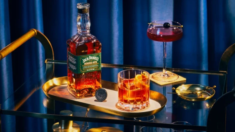 Bottle of Jack Daniel's Bonded Rye and two cocktails on a gold bar cart against a dark blue curtain backdrop.