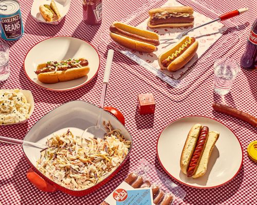 Overhead shot of multiple hot dogs on a red and white checkered tablecloth, with bowls of sides and branded condiments.