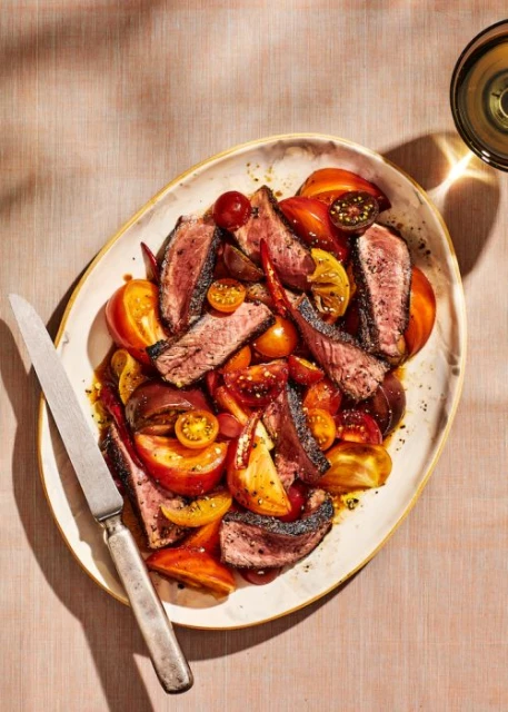 Overhead view of seared steak slices and heirloom tomatoes served on a platter, next to a silver knife and a glass of wine.