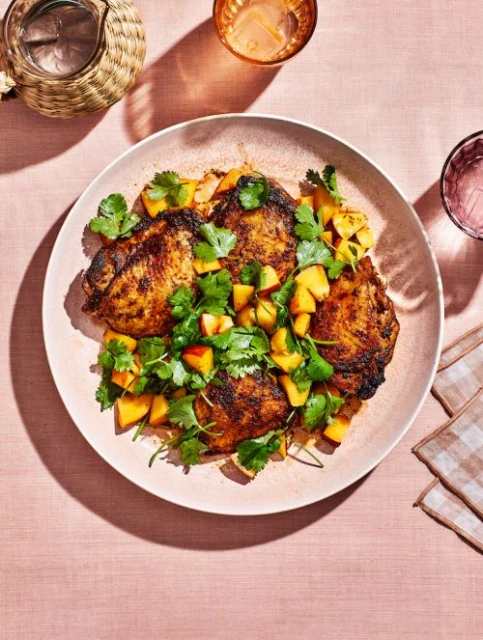 Overhead shot of a plate of grilled chicken thighs with peach salsa and cilantro garnish on a pink tablecloth next to glassware.