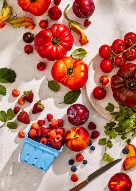 Overhead shot of a variety of fresh summer fruits and vegetables including tomatoes, plums, peaches, raspberries, strawberries, and basil.