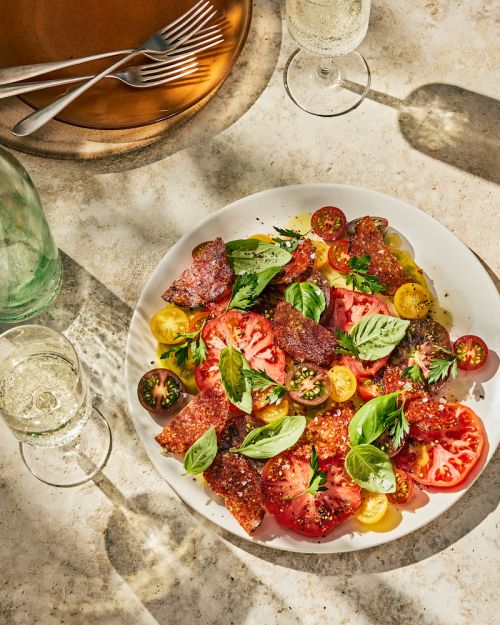 Overhead shot of a tomato and basil salad with crispy prosciutto on a white plate, accompanied by champagne flutes and plates.
