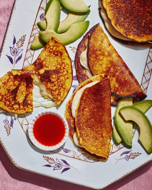 Overhead shot of corn arepas with avocado slices and red dipping sauce on a patterned plate against a pink fabric surface.