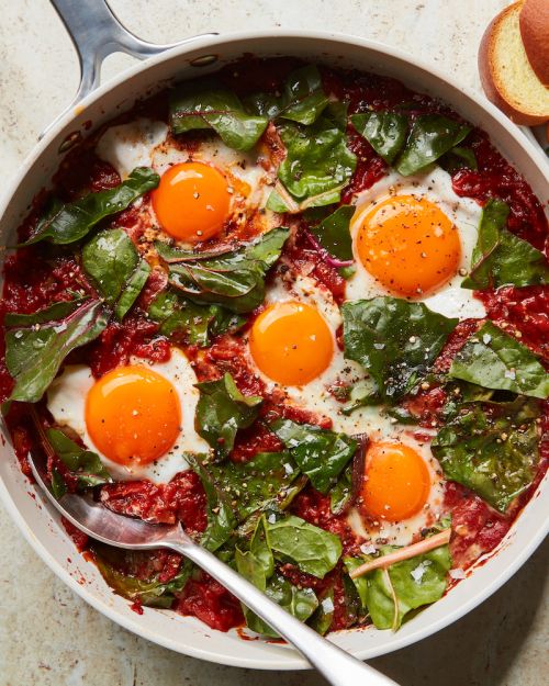 Overhead shot of shakshuka with five fried eggs, tomato sauce, and spinach in a pan, topped with bread slices for a hearty breakfast.