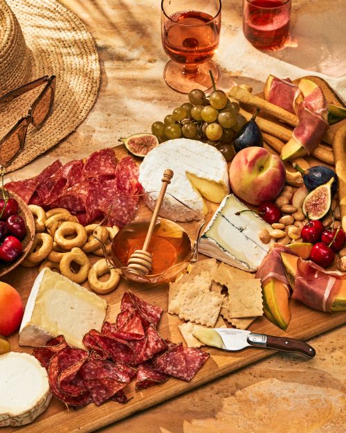 Overhead shot of charcuterie board with cheeses, meats, fruit, and nuts, with glasses of rosé and a straw hat in the background.