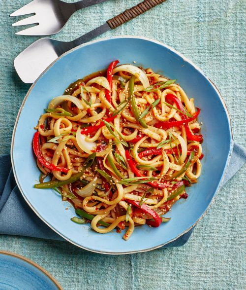Overhead shot of noodles with red bell peppers and green onions in a blue ceramic bowl on a blue tablecloth, featuring serving utensils.