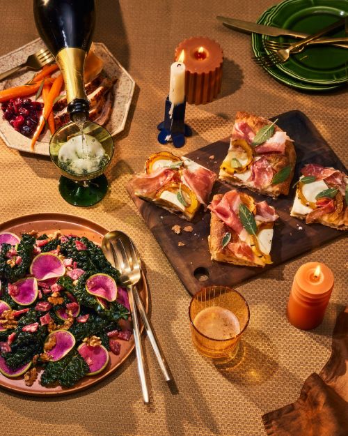 Overhead shot of a Thanksgiving feast including champagne pouring into glass, turkey with sides, salad, and pizza on a neutral tablecloth.