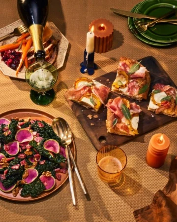 Overhead shot of a Thanksgiving feast including champagne pouring into glass, turkey with sides, salad, and pizza on a neutral tablecloth.