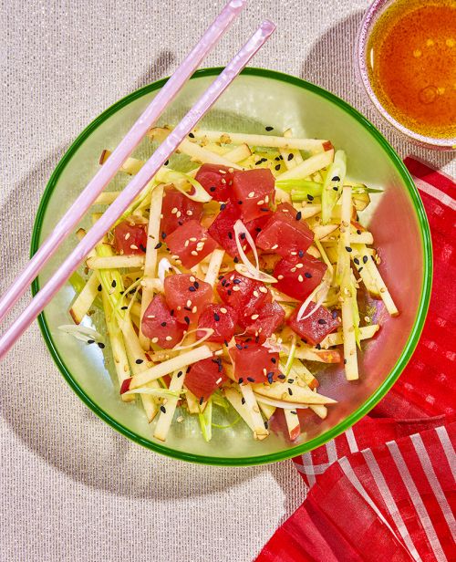 Overhead shot of a vibrant tuna salad with tuna cubes, sesame seeds, and long chopsticks resting on a green bowl and red cloth.