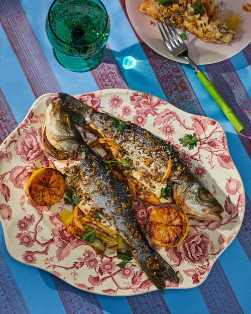 Overhead shot of two cooked fish garnished with lemon slices and herbs on a patterned plate with striped backdrop. Food photography.