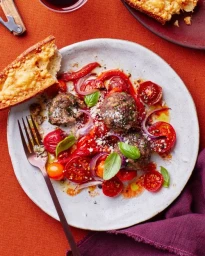 Overhead food shot of meatballs with roasted cherry tomatoes, red onions, and basil on a speckled white plate with a fork and toasted bread.