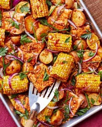 Overhead shot of a metal baking sheet filled with seasoned corn, roasted potatoes, chicken pieces, red onion slices, and fresh herbs.