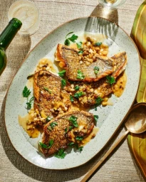 Pan-seared fish fillets with herb and nut garnish on a speckled ceramic oval platter over a textured tablecloth.
