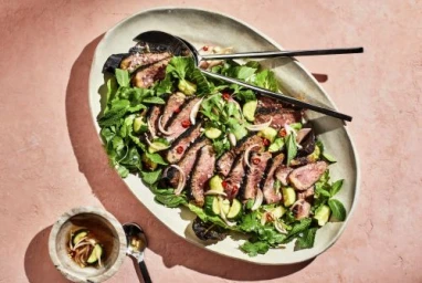 Sliced grilled steak salad with fresh herbs and cucumber served on a large oval ceramic platter on pink textured surface