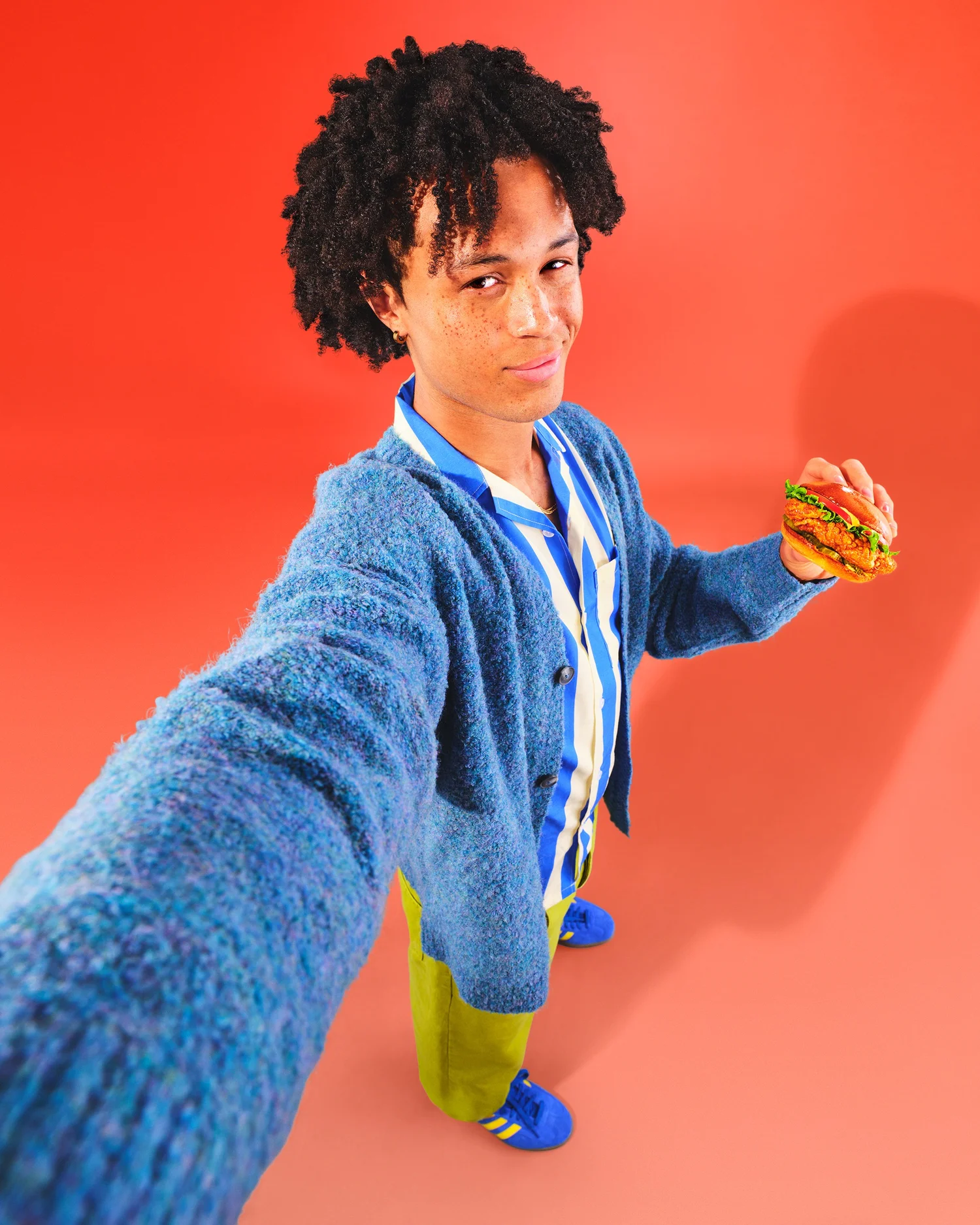 Young man takes a selfie in a blue cardigan and green pants, holding a chicken sandwich against a gradient orange background.