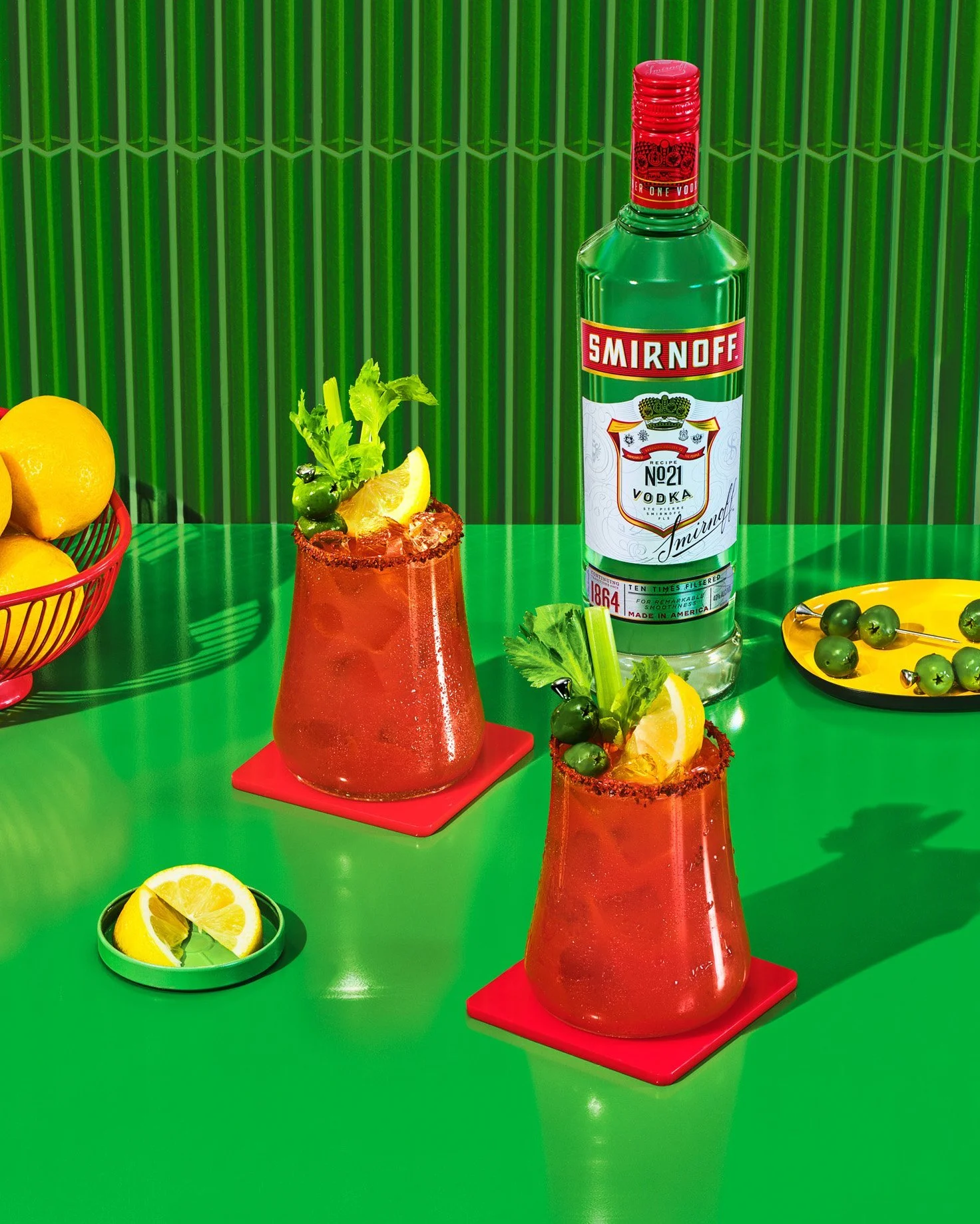 Two bloody mary cocktails with celery and lemon wedges, served on a green table with lemons, olives, and a bottle of Smirnoff vodka.