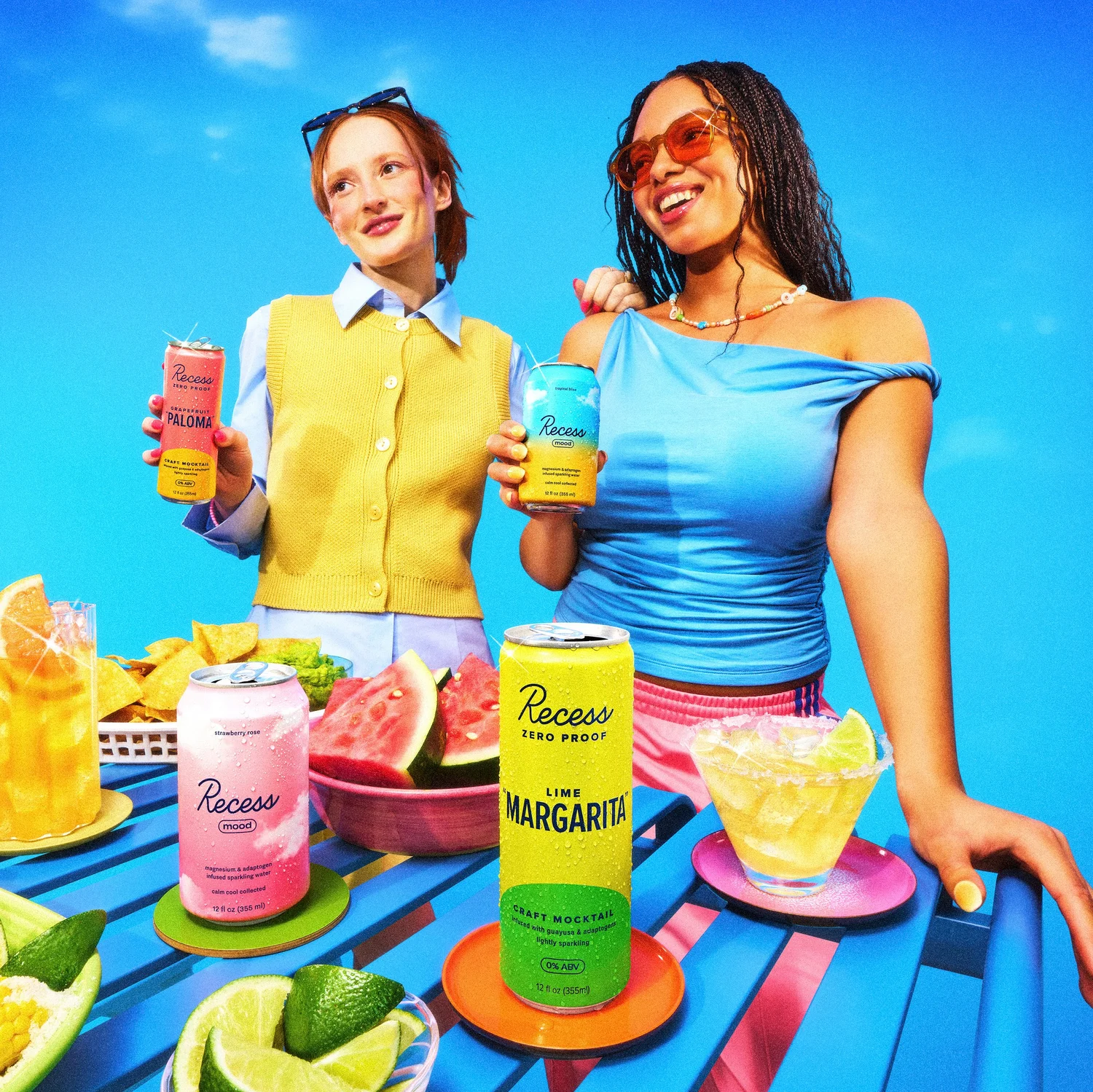 Two women hold Recess beverages at an outdoor table with assorted appetizers against a vibrant blue sky on a sunny day.