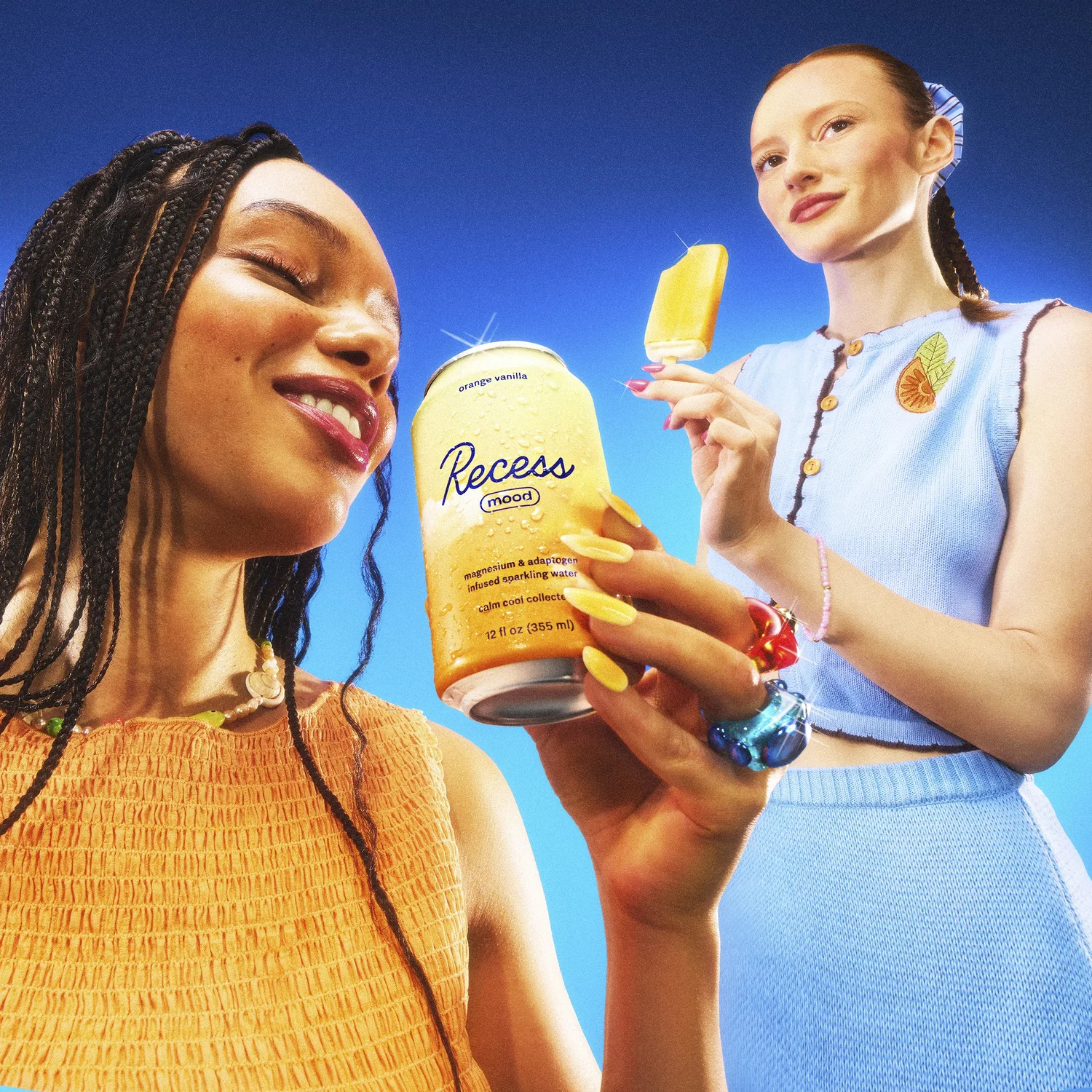 Two women pose with a can of Recess mood drink and a popsicle against a blue backdrop in a vibrant and playful advertising shot.