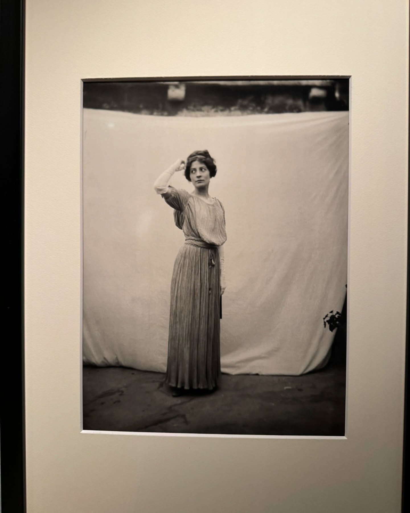 Monochrome studio shot of an adult female model posing in long pleated gown with hand on head, against white backdrop.