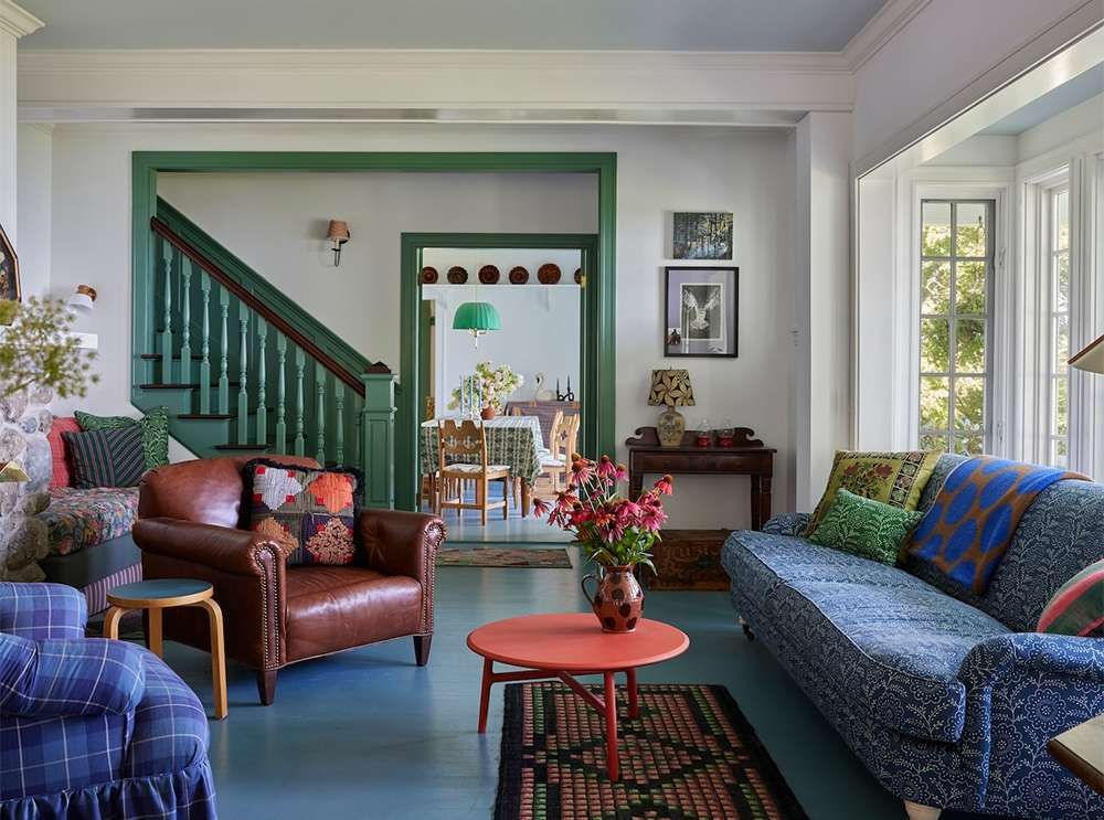 Living room interior with a blue patterned couch, a brown leather armchair, a red table with flowers, and a green staircase and trim.