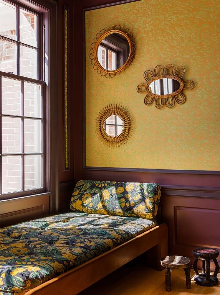 Interior corner featuring a daybed with floral upholstery, three decorative mirrors on a yellow patterned wall, and small wooden stools.