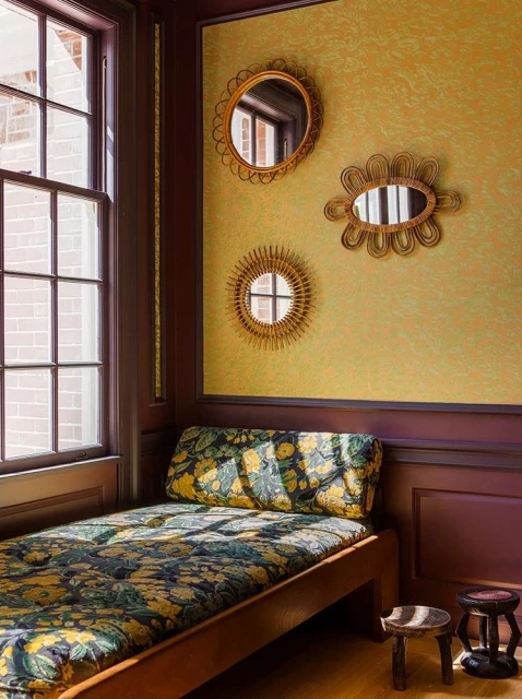 Interior corner featuring a daybed with floral upholstery, three decorative mirrors on a yellow patterned wall, and small wooden stools.