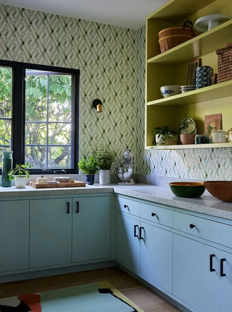 Bright, contemporary kitchen interior with sage green wallpaper, light blue cabinets, and a window overlooking greenery.
