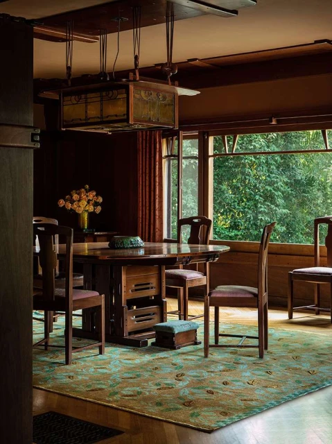 Interior dining room featuring a wood table and chairs, rectangular pendant light, green patterned rug, and a view of lush greenery.