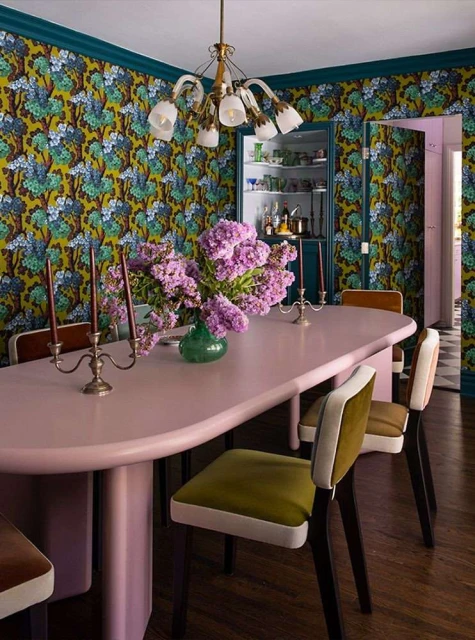 Interior design featuring a pink dining table with lilac flowers, surrounded by chairs in a dining room with patterned wallpaper.