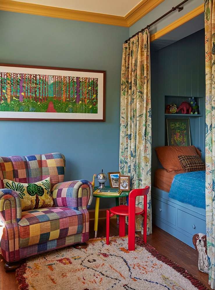 A colorful children's bedroom featuring a patchwork armchair, a small red chair, a daybed nook with patterned curtains, and framed artwork.
