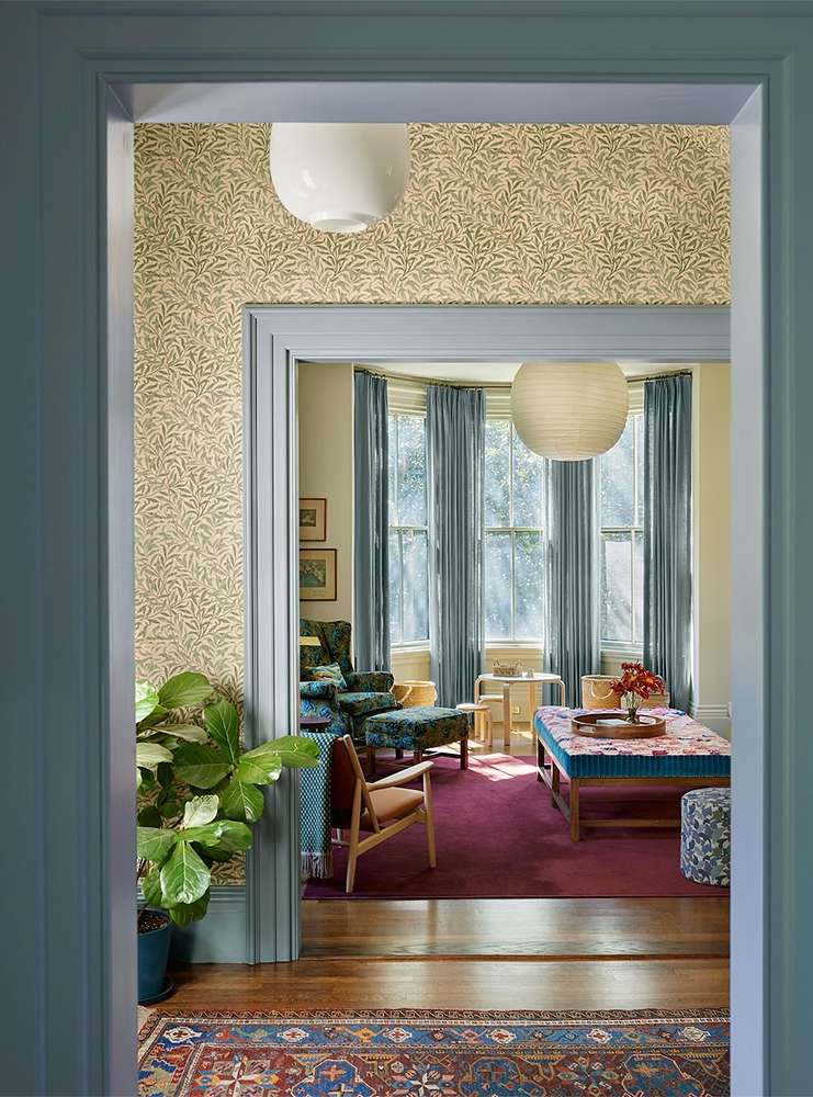 Well-lit living room seen through a doorway, featuring a patterned rug, upholstered furniture, hardwood floors, and natural light.