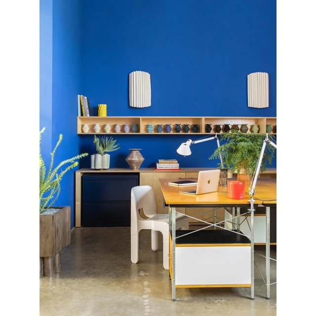 Blue-walled home office with a wooden desk, laptop, white chair, and various decorative items on a shelf and desk surface.