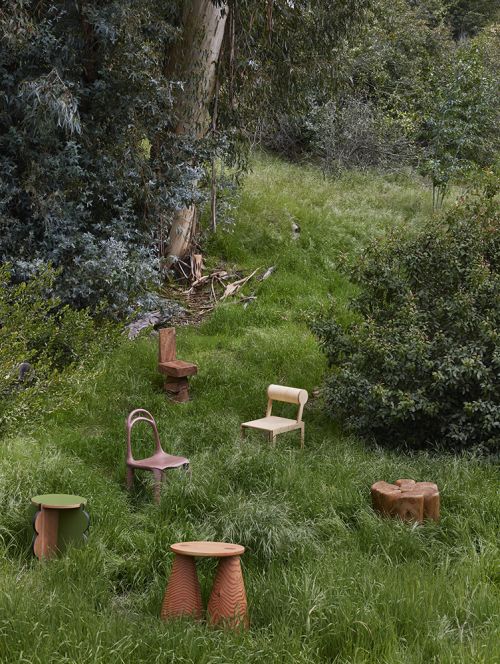 A collection of wooden chairs and stools are arranged on a grassy hill, surrounded by trees in a natural outdoor setting. 