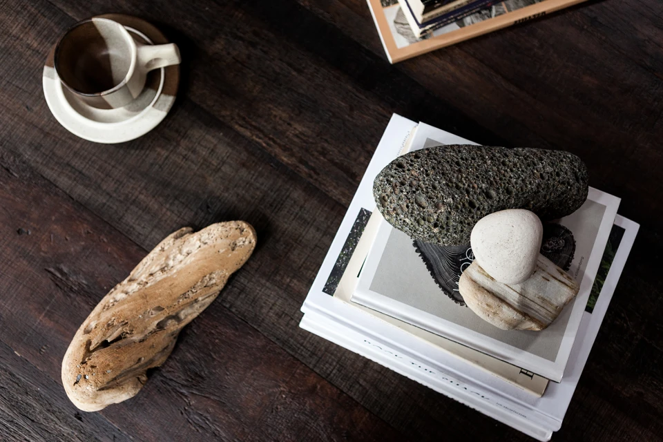 Still life arrangement on a dark wood table featuring stacked books, smooth stones, a unique piece of driftwood, and a two-tone coffee cup.