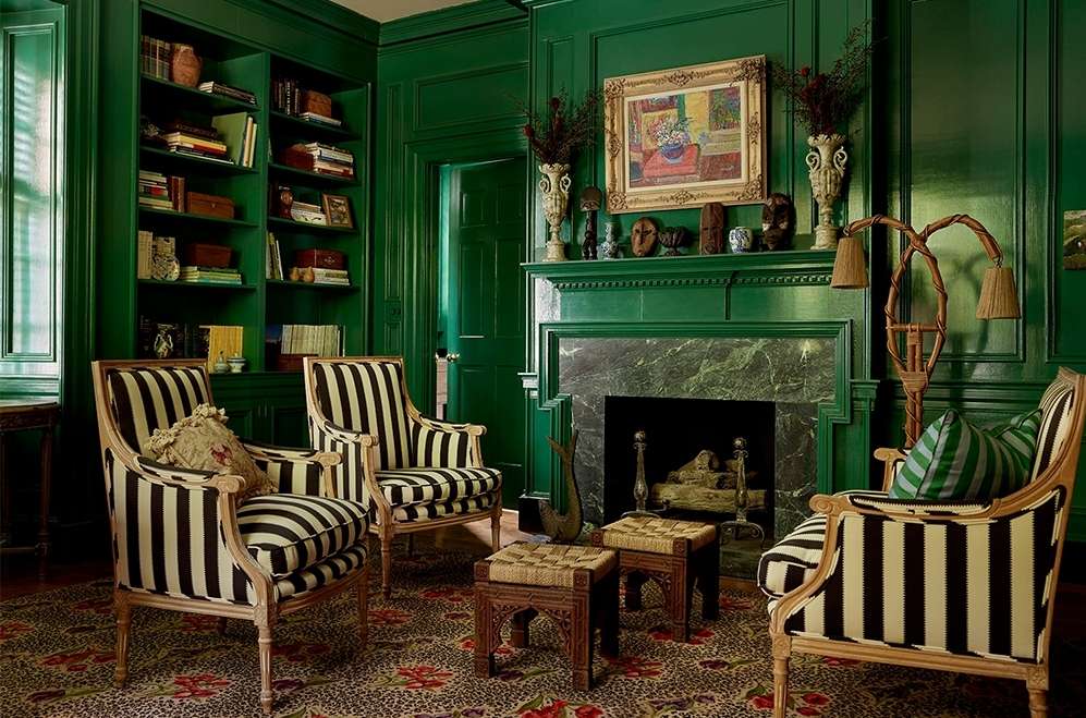 Living room interior with four striped chairs around a patterned rug, fireplace with mantle, dark green walls, and built-in bookcase.