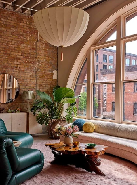 Living room interior with an exposed brick wall, modern green armchairs, a wooden coffee table, and large arched windows.