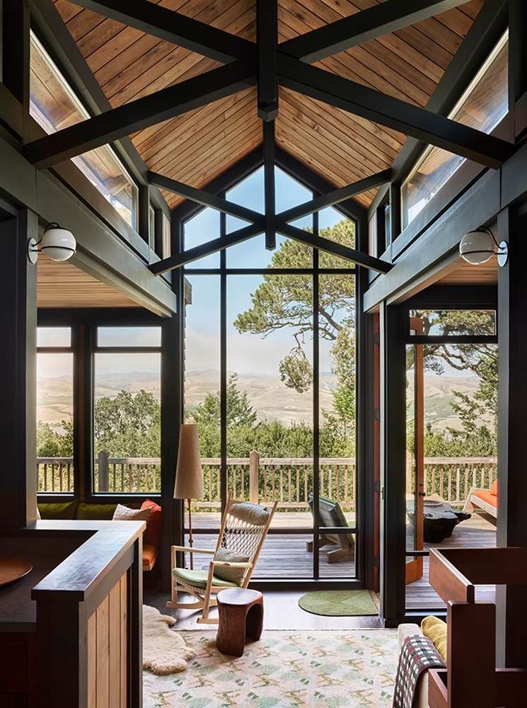 Interior view of a modern cabin with a pitched wood ceiling, large windows, a rocking chair, and a view of a wooded landscape.