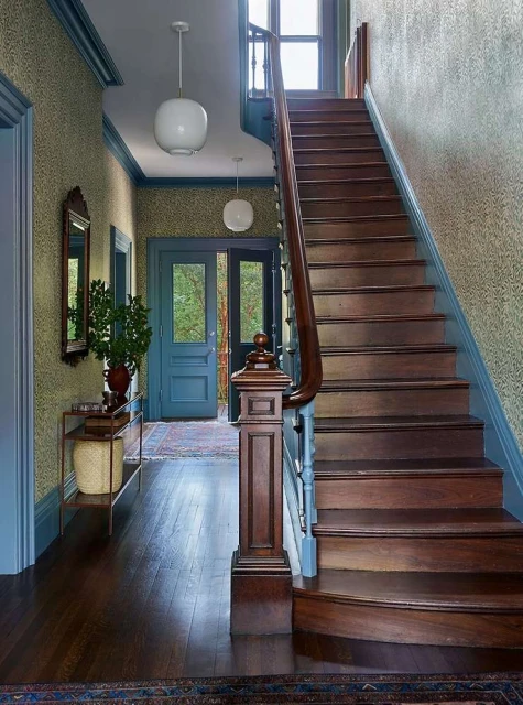Interior view of a home's foyer with a wooden staircase, blue trim, patterned wallpaper, pendant lights, and an open doorway.