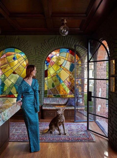 Woman in teal gown poses next to dog in interior with stained-glass windows, wood floors, and brick accents. Indoor setting.