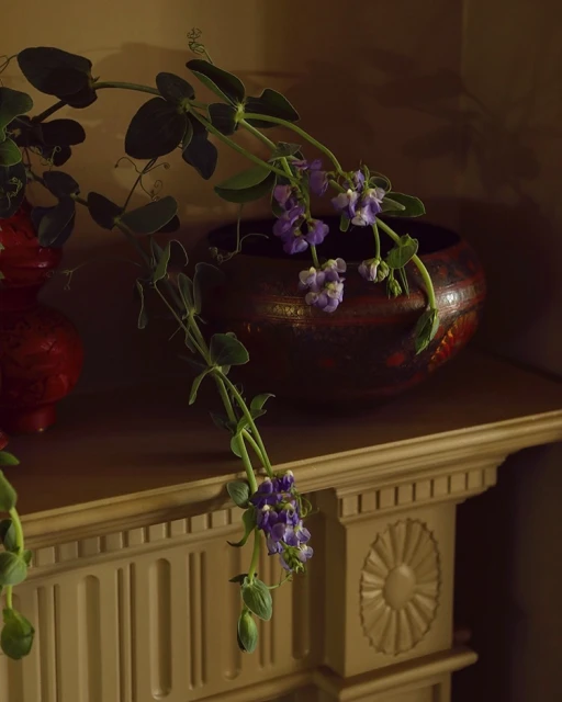 Floral arrangement featuring purple flowers and green leaves in a decorative red bowl, set on a beige shelf with molded detail.