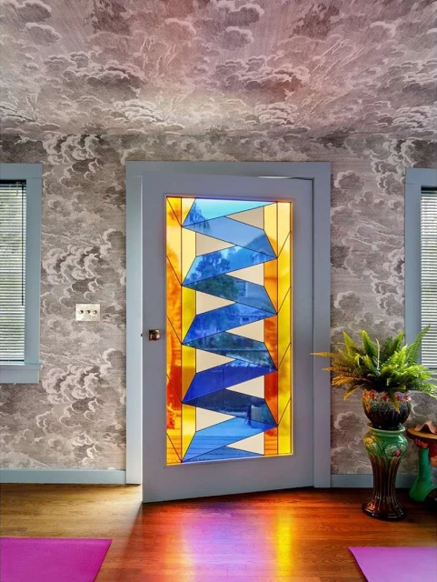 Stained glass door with geometric pattern in modern interior with cloud wallpaper, wood floor and fern. Sunlight casts rainbow effect.