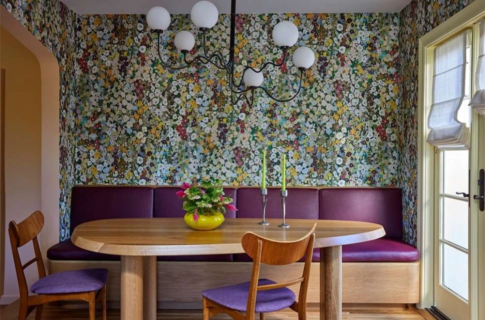 Interior dining space featuring a light wood table, seating area with purple cushions, and a floral wallpaper backdrop.