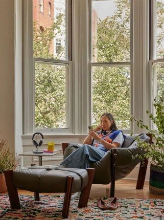 Woman seated in a leather armchair using a smartphone in a living room with large windows by photographer Laure Joliet.