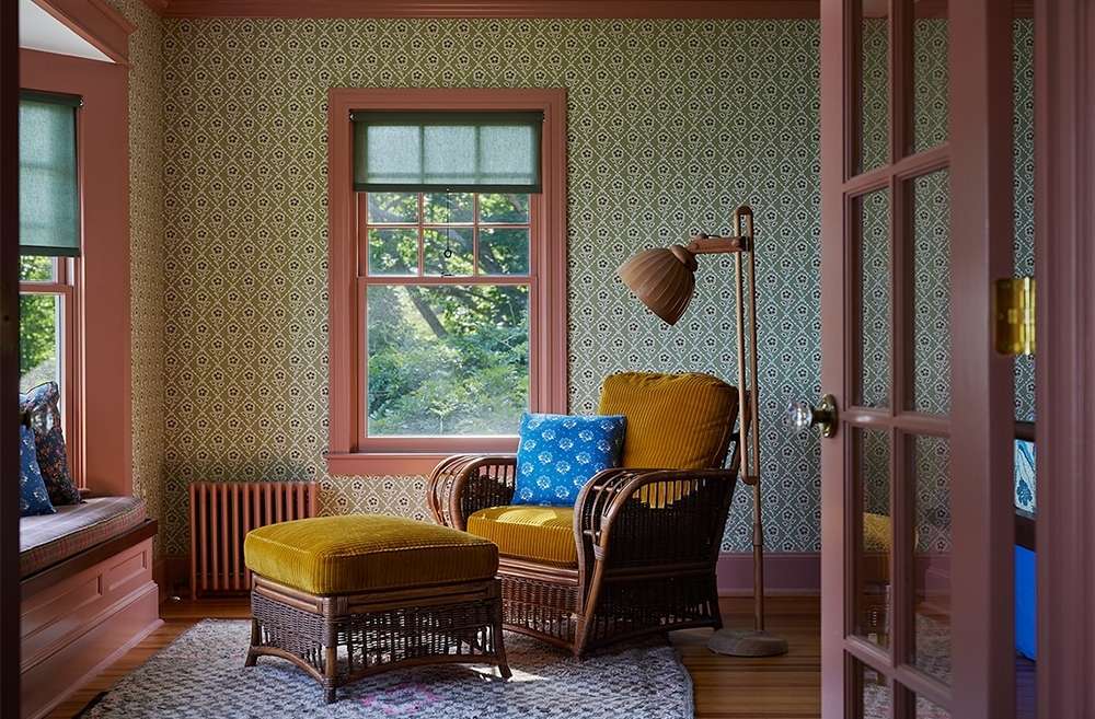 Cozy living room interior with rattan armchair, ottoman, window with green view, patterned wallpaper and pink trim in natural daylight.