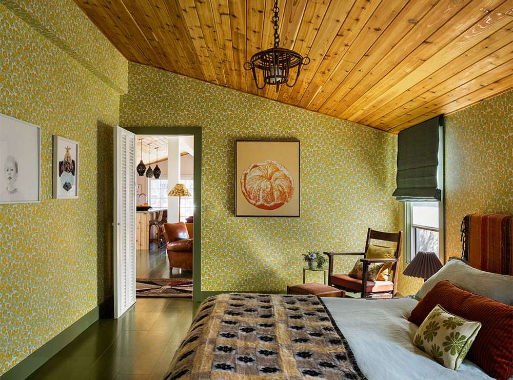 Bedroom interior featuring a bed with patterned blanket, armchair, and floral wallpaper, illuminated by natural light.
