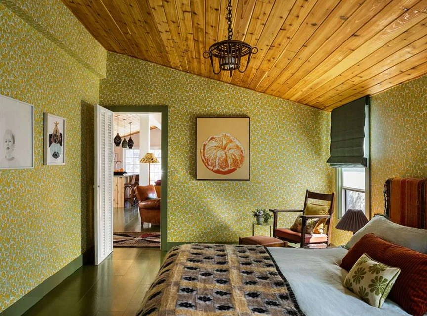 Bedroom interior featuring a bed with patterned blanket, armchair, and floral wallpaper, illuminated by natural light.