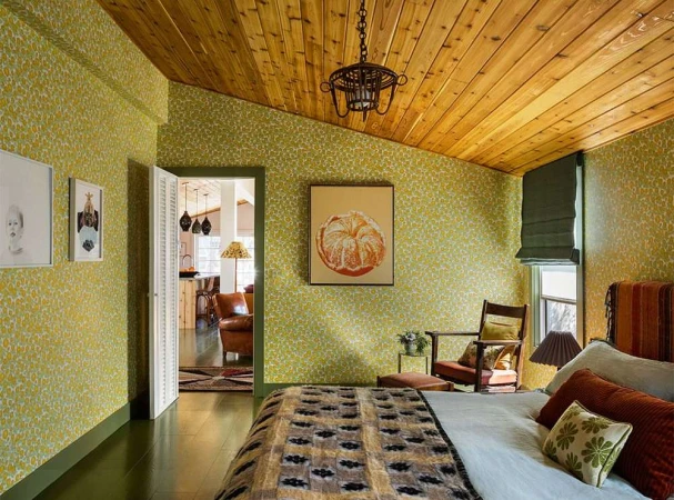 Bedroom interior featuring a bed with patterned blanket, armchair, and floral wallpaper, illuminated by natural light.