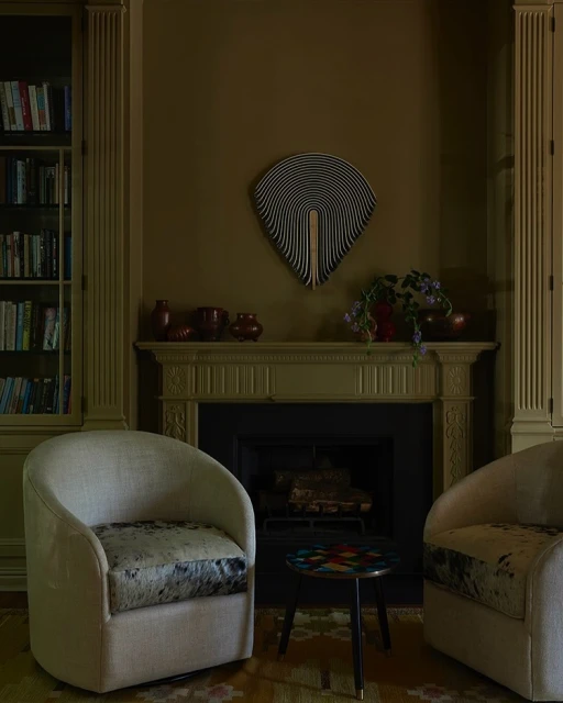 Interior of a living room with two armchairs, a small table, fireplace and mantel, a decorative wall art, bookshelves, and dark green walls.